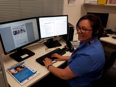 A woman sits in front of two computer monitors. She wears a headset. 