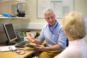 A male doctor sits face to face with an elderly woman. They are in the doctors office . The focus is on the doctor who sits at his desk holding a digital tablet and smiling at the woman. The woman face cannot be seen.