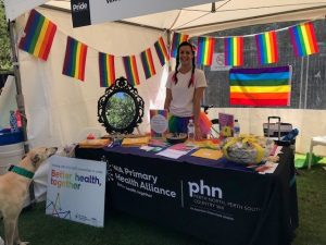 A woman stands at a stall. The LGBTI flag covers teh stall. 